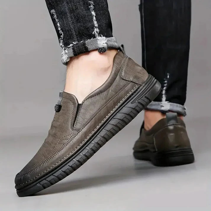 Brown slip-on shoes worn with black jeans on a neutral background